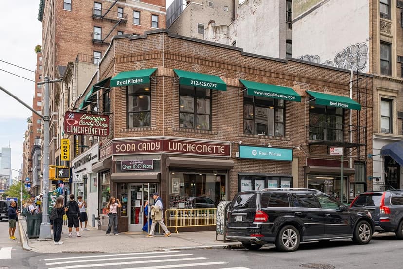 Exterior of Lexington Candy Shop, a corner building with green awnings, on a New York City street with pedestrians and parked cars.