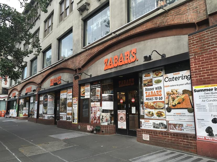 Zabar's grocery and deli storefront on a city street with a sidewalk, featuring multiple display windows and brick arches.