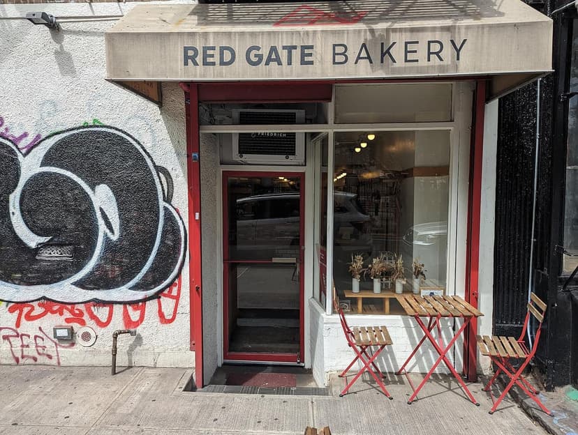 Exterior view of Red Gate Bakery storefront with a red door, a display window, and two red outdoor tables with chairs on the sidewalk.