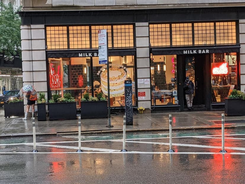 Exterior of a Milk Bar storefront on a rainy day. A person with an umbrella stands left, another person in the doorway right.