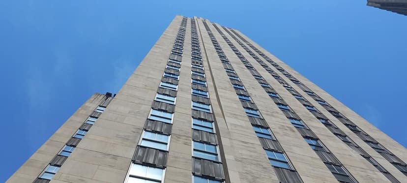 A tall skyscraper with stone facade and numerous windows, seen from a low angle against a clear blue sky.