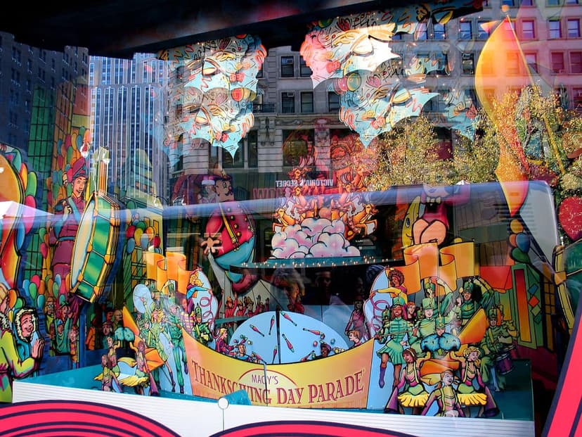 A colorful Macy's Thanksgiving Day Parade window display featuring cartoon characters, drums, and a banner. City buildings are reflected in the