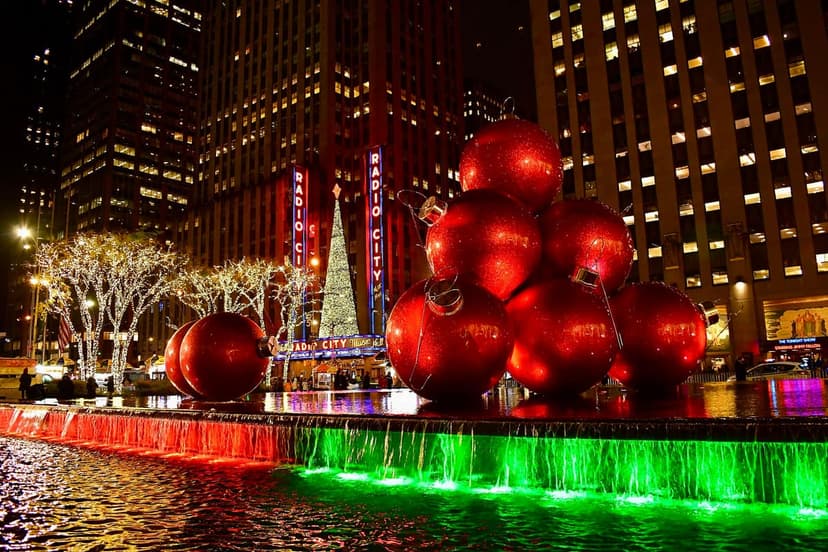 Radio City Music Hall exterior at night with holiday decorations, including a Christmas tree, large red ornaments, and a fountain with red and