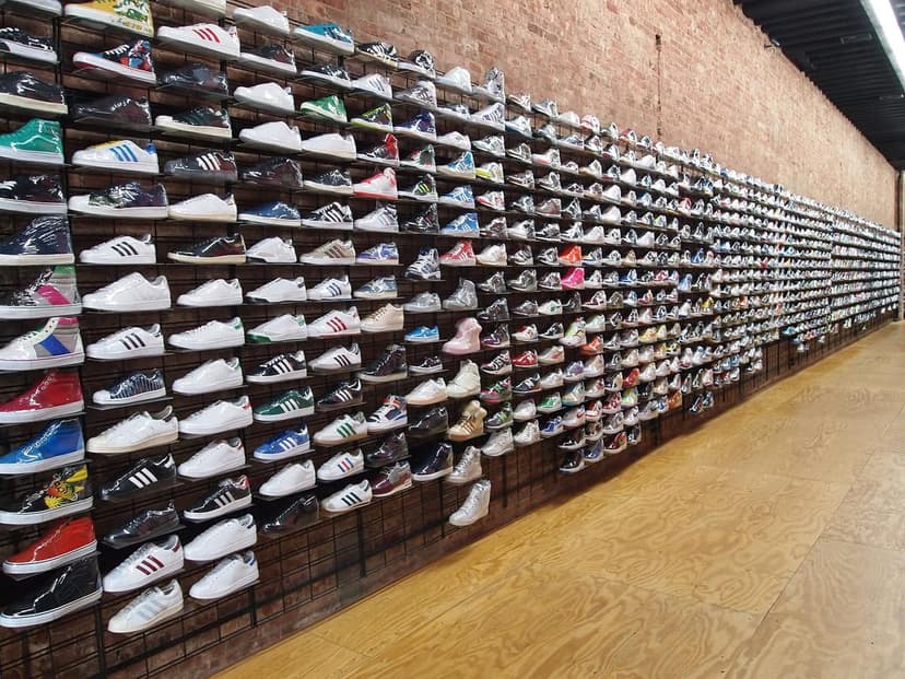 A long wall inside a sneaker store is covered with hundreds of pairs of shoes displayed on shelves against a brick wall.