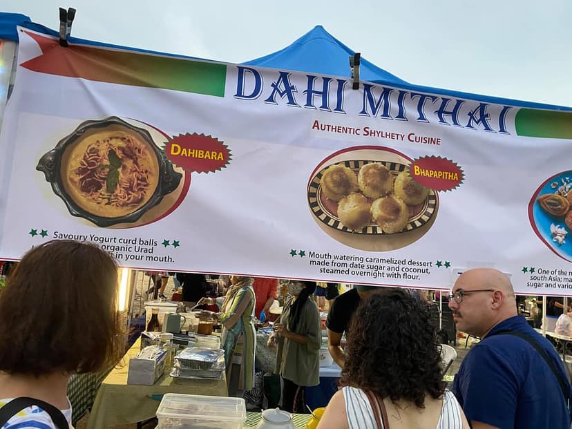 A food stall at an outdoor market with a banner advertising Dahi Mithai Authentic Shylhety Cuisine.