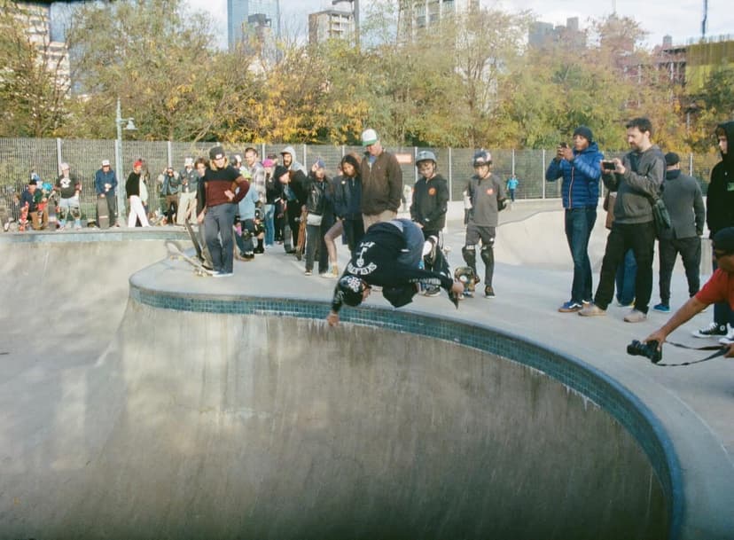 A skateboarder performs an air trick in a concrete skatepark bowl, surrounded by spectators watching the event.