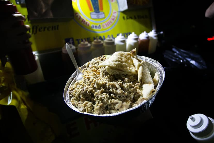 A foil container holds chicken and rice with pita bread and a spoon. A food cart banner and sauce bottles are in the background.