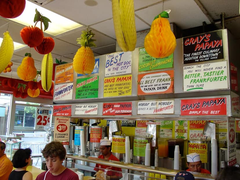 Interior of Gray's Papaya with customers, staff, and signs advertising hot dogs and papaya drinks. Paper fruit decorations hang.