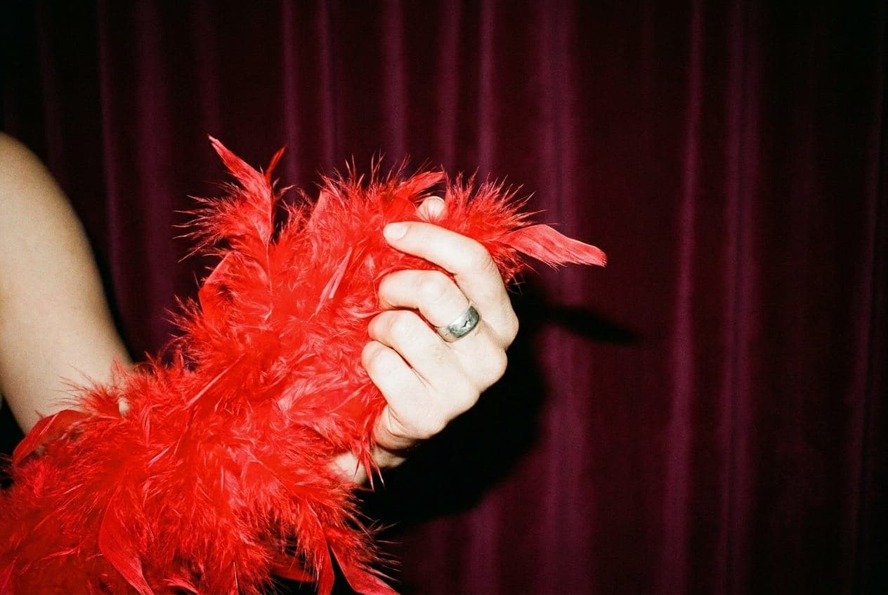 A performer's hand holding a bright red feather boa against a velvet curtain