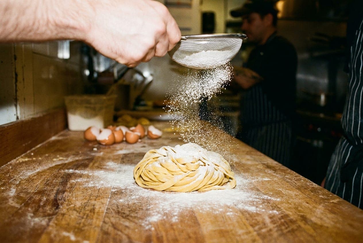 Hands dusting flour over fresh pasta dough on a wooden board.