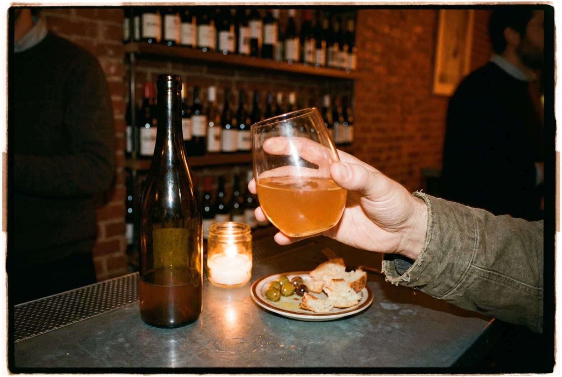 A hand tilting a glass of cloudy orange wine at a candlelit zinc bar, with a chalkboard wine list and bottles blurred in the background