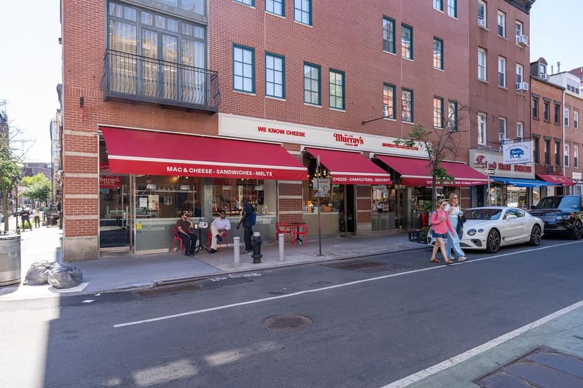 Exterior of Murray's Cheese shop with red awnings on a brick building. People are seated at outdoor tables and walking on the sidewalk.