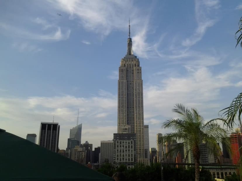 The Empire State Building stands tall against a blue sky with white clouds, viewed from a rooftop bar with palm trees and other city
