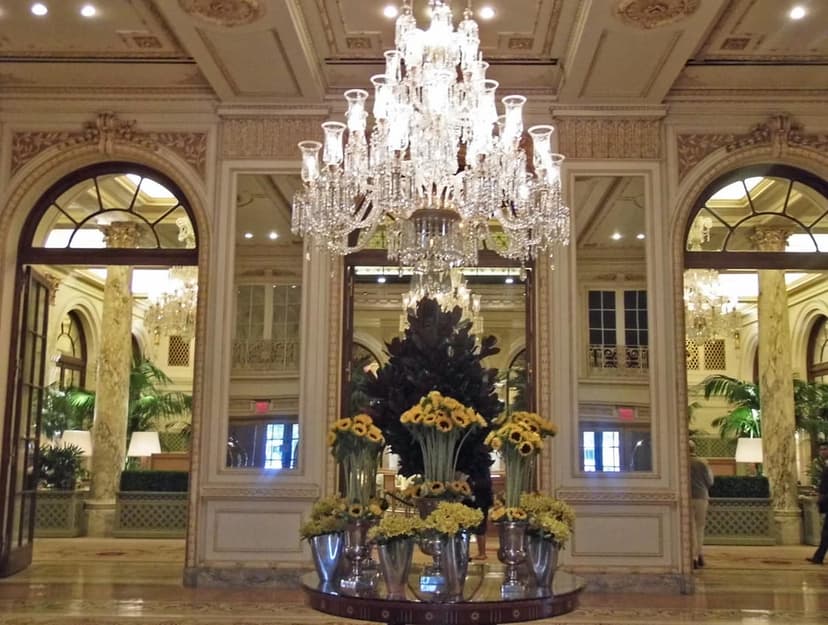 Grand hotel lobby with a large crystal chandelier, ornate ceilings, arched doorways, mirrors, and yellow floral arrangements.