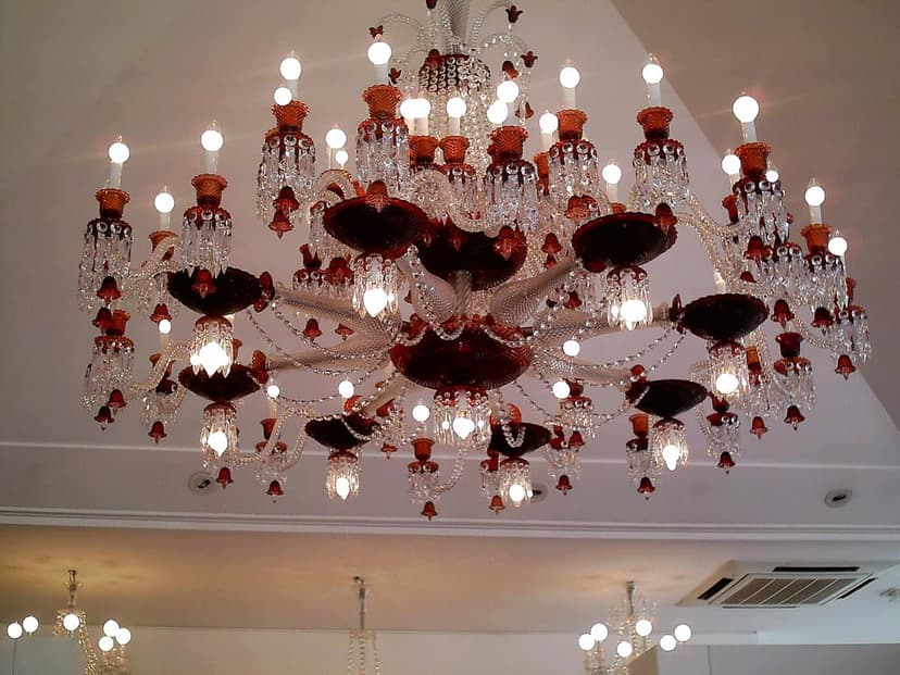 A large, ornate crystal chandelier with red glass accents hangs from a white ceiling, illuminated by many small bulbs.