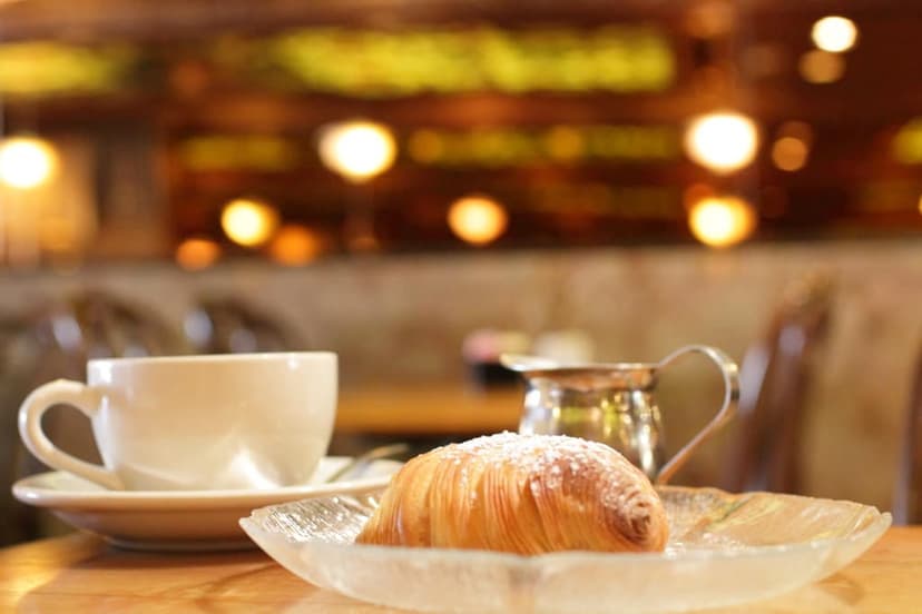 A white coffee cup on a saucer, a sfogliatella pastry dusted with powdered sugar on a clear glass plate, and a silver creamer on a table in a