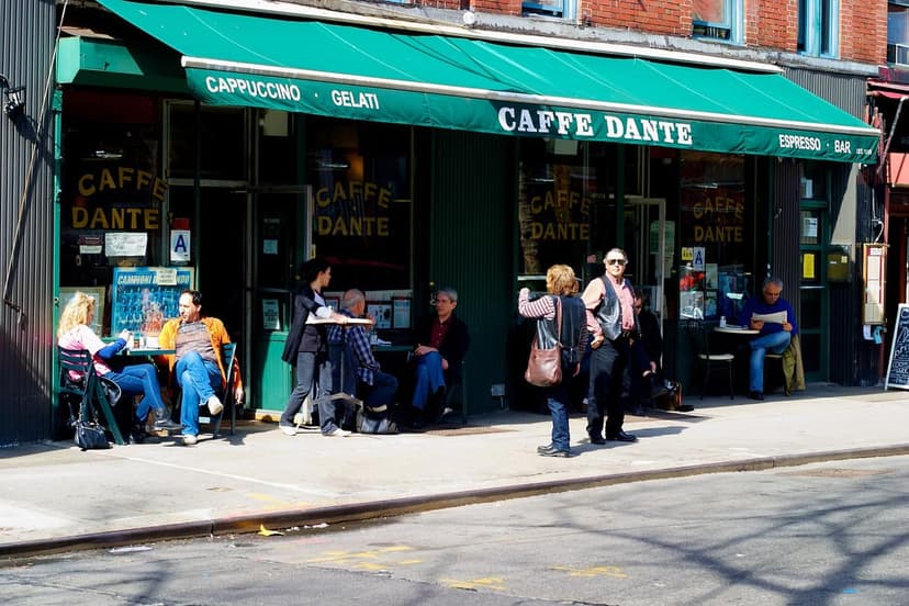 The exterior of Caffe Dante on a sunny day, with a green awning and several patrons seated at outdoor tables on the sidewalk.