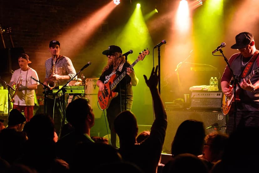 A band performs on a stage under yellow and orange lights, with musicians playing saxophone, trumpet, bass, and electric guitar to a