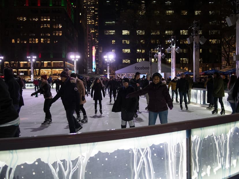 People ice skating on an outdoor rink at night, with illuminated city buildings and a 'facebook' tent in the background.