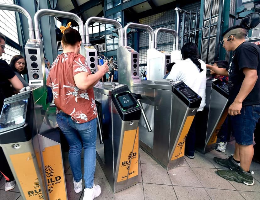 People are passing through subway turnstiles with OMNY readers. One person is tapping a card on a reader that displays 'GO'.