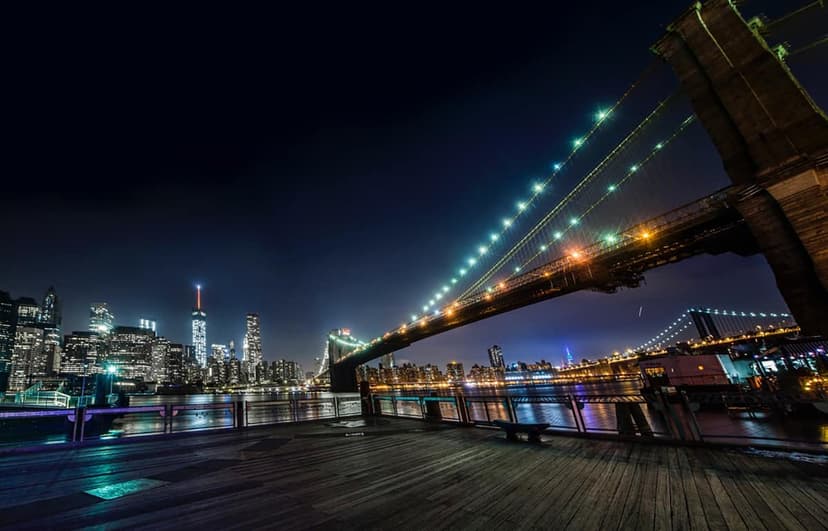 Nighttime view from a wooden pier in Brooklyn, showing the illuminated Brooklyn Bridge, the Manhattan skyline, and the East River.
