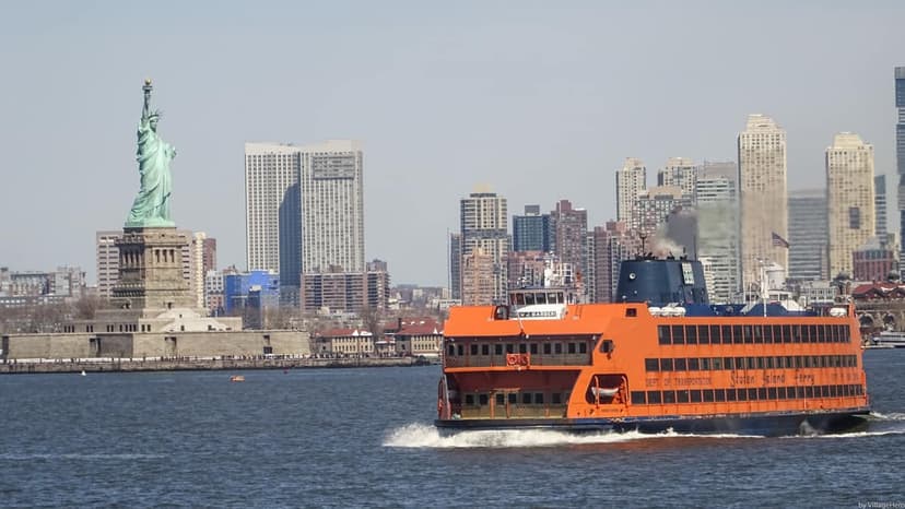An orange Staten Island Ferry sails past the Statue of Liberty with the New York City skyline in the background on a clear day.