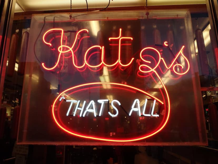 A red neon sign for Katz's Delicatessen reads 'Katz's' in script and 'THAT'S ALL' in white letters within a red oval.