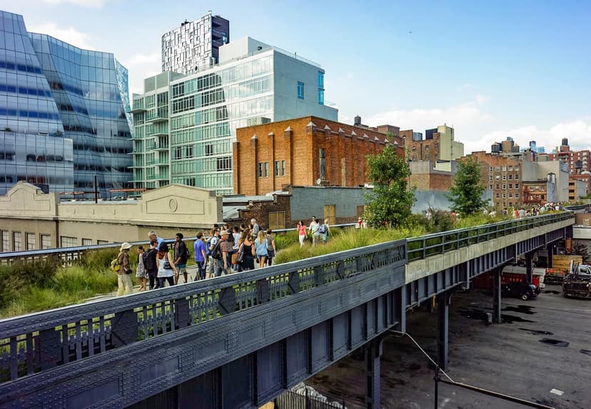 People walk on the High Line park, an elevated green space, with modern glass buildings and traditional brick structures in the