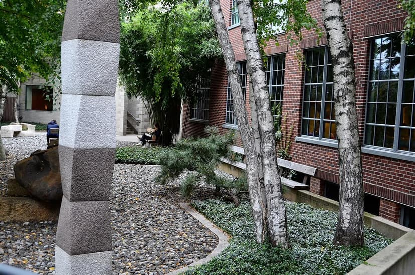 A garden with a tall, multi-toned stone sculpture, two birch trees, and a brick building with windows. People sit on benches.