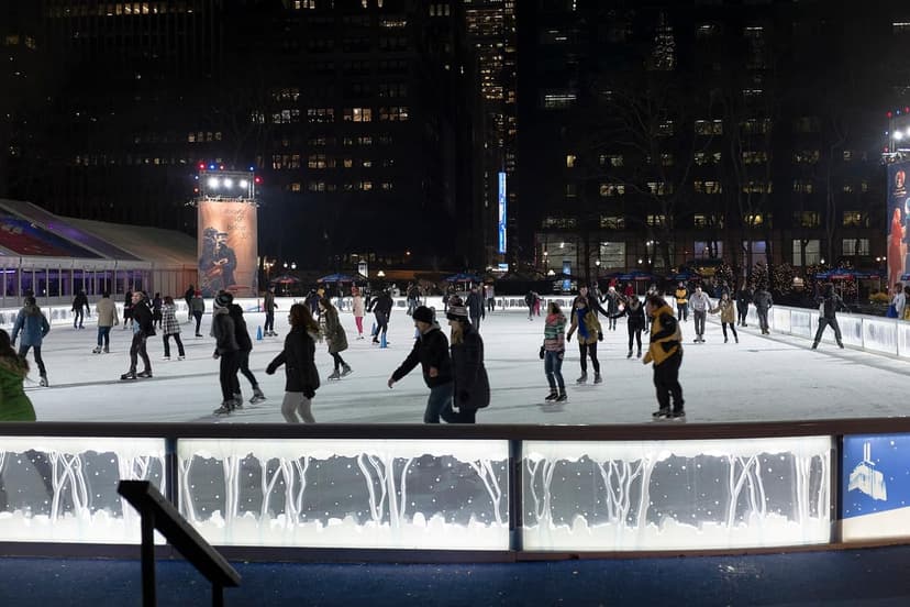 People ice skating on an outdoor rink at night in a city park, with tall buildings in the background.