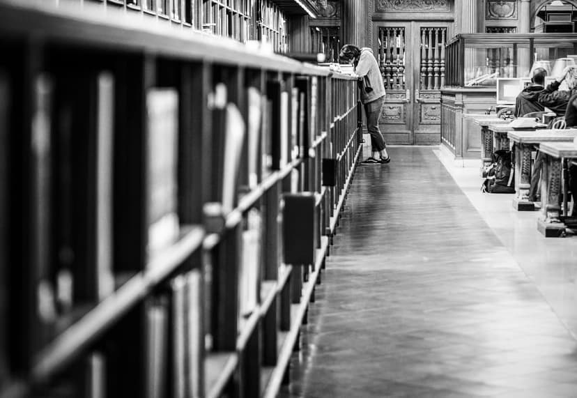 Black and white photo of a person leaning against a tall bookshelf, reading in a grand library hall with ornate architecture.
