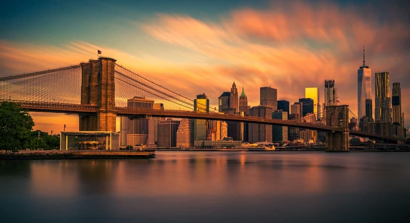 The Brooklyn Bridge spans a body of water with the Manhattan skyline visible under a colorful sunrise sky.