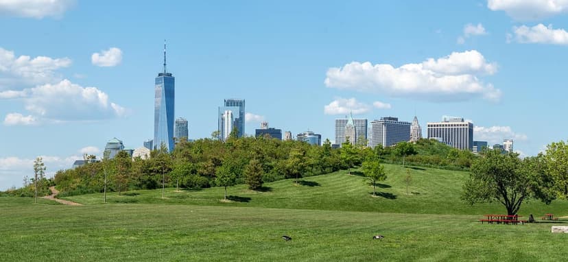 A panoramic view of a green park with rolling hills and trees, with the Manhattan skyline and One World Trade Center.