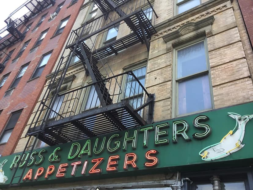 Exterior view of Russ & Daughters Appetizers sign with neon letters on a green background, below a brick building with fire escapes.