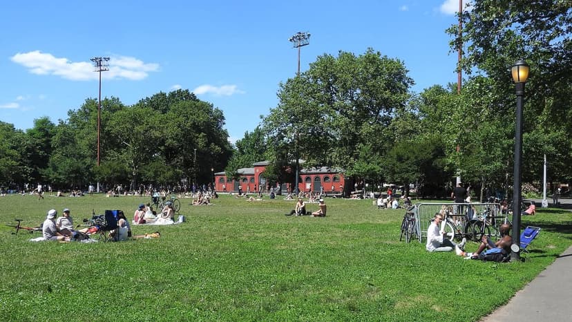 A wide view of a sunny green park with numerous people relaxing on the grass. Trees, a red brick building, and tall light poles are