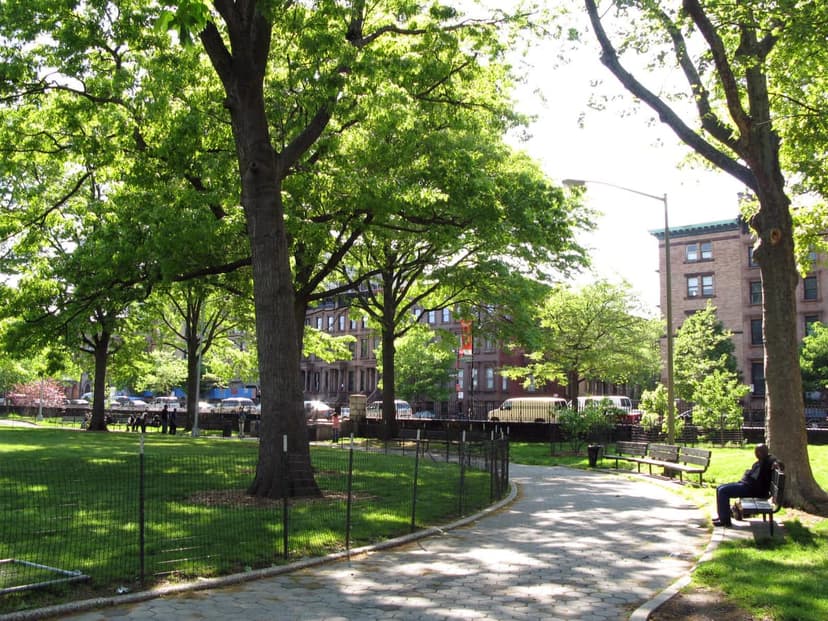 A paved path winds through a green park with many trees, leading past a grassy area enclosed by a low fence.