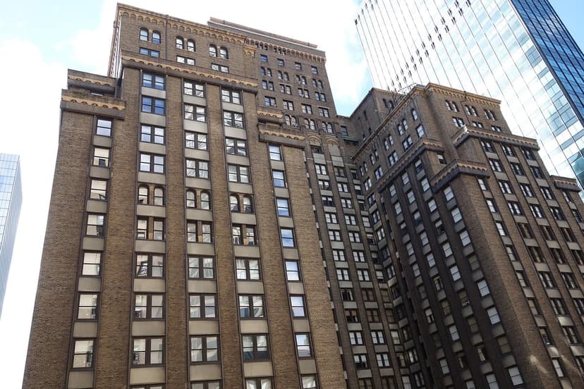 A tall, brown brick building with numerous windows, viewed from below, with a modern glass skyscraper partially visible.