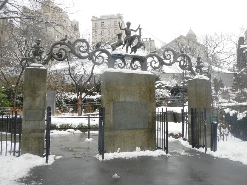 The Lehman Gates of the Central Park Zoo are covered in snow, featuring ornate scrollwork and sculptures of children and animals.