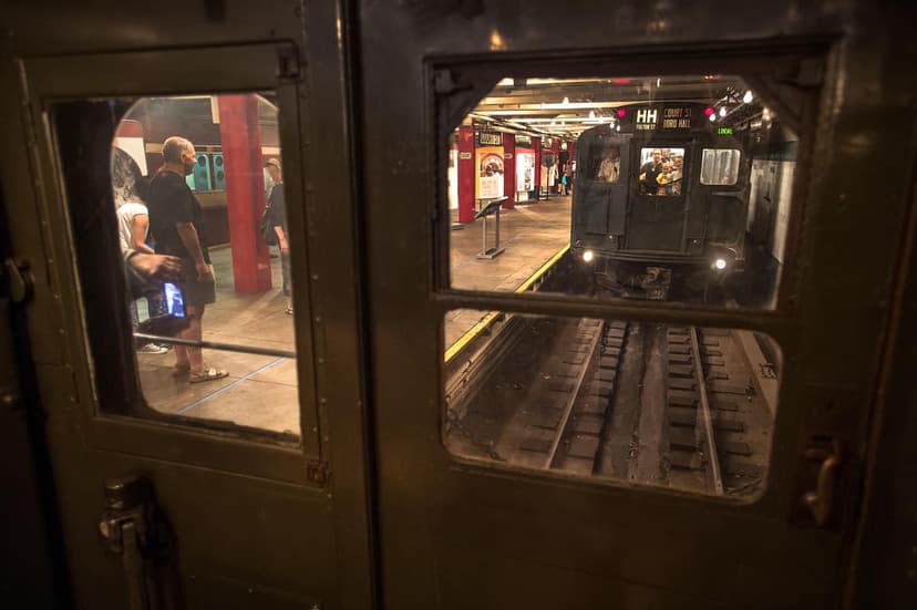 View from inside a subway car looking through a window at another subway car on tracks and a platform with red pillars.
