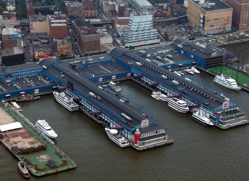 Aerial view of Chelsea Piers, a large complex of blue buildings on a waterfront with boats docked. City buildings are in the background.