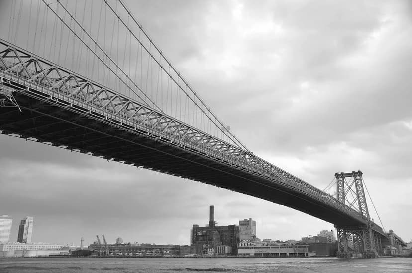 A black and white low-angle view of the Williamsburg Bridge over the East River, with the Domino Sugar Refinery building on the Brooklyn