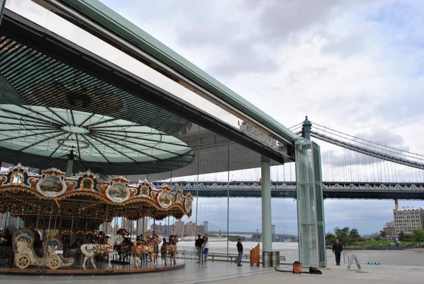 Jane's Carousel inside its glass pavilion with the Manhattan Bridge and East River in the background under a cloudy sky.