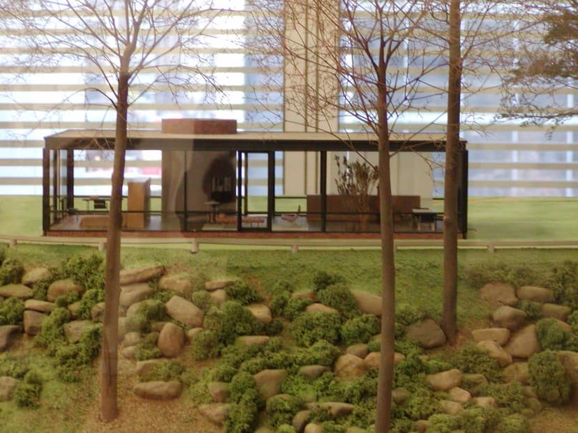 An architectural model of Philip Johnson's Glass House, with a transparent structure, miniature trees, and rocks, displayed inside a