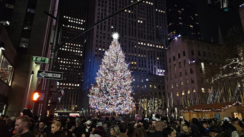 A brightly lit Christmas tree stands tall in Rockefeller Center at night, surrounded by tall buildings and a large crowd of people.
