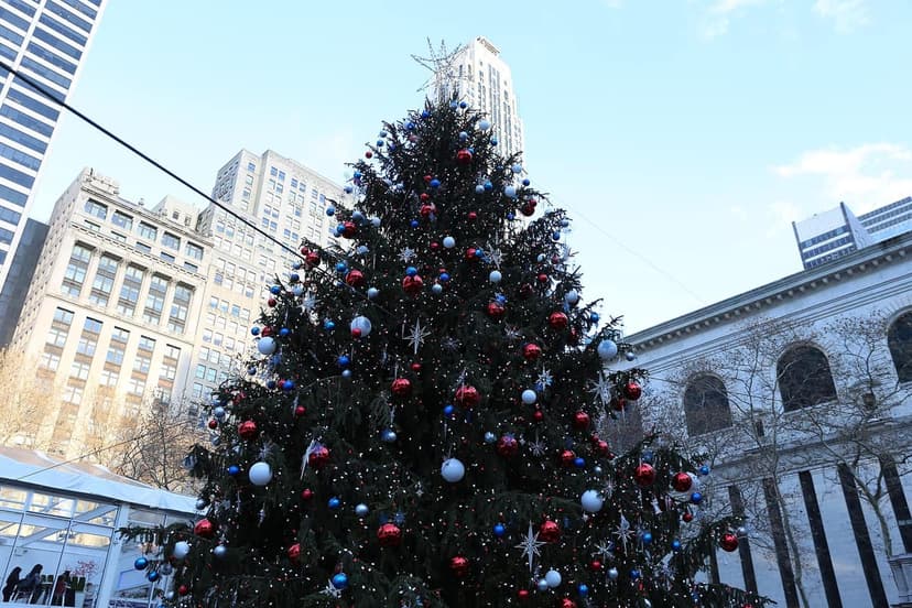 A large outdoor Christmas tree adorned with red, blue, and white ornaments and lights stands in a city park. Buildings are in the