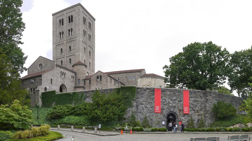 Exterior view of The Met Cloisters, a medieval-style stone building with a tall tower, surrounded by trees and a stone wall with red