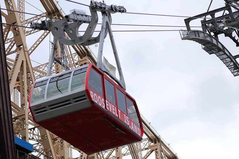 A red and grey Roosevelt Island tram car is suspended on cables, with 'ROOSEVELT ISLAND' written on its side. A bridge structure is in the