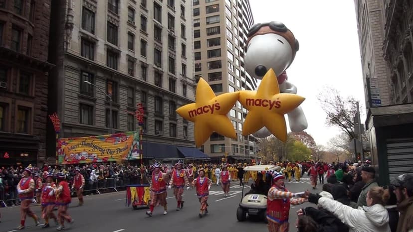 A large Snoopy balloon and two yellow Macy's star balloons float above a street parade with costumed participants and spectators.