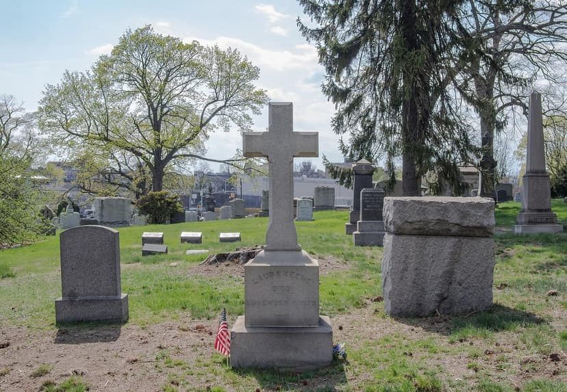 A cross-shaped gravestone for Laura Keene in Green-Wood Cemetery, with other gravestones and trees in the background.