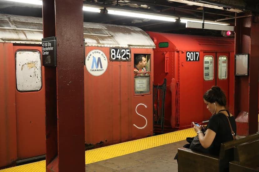 Red subway train cars are at a station platform. A man looks from the driver's window of car 8425. A woman sits on a bench looking at her